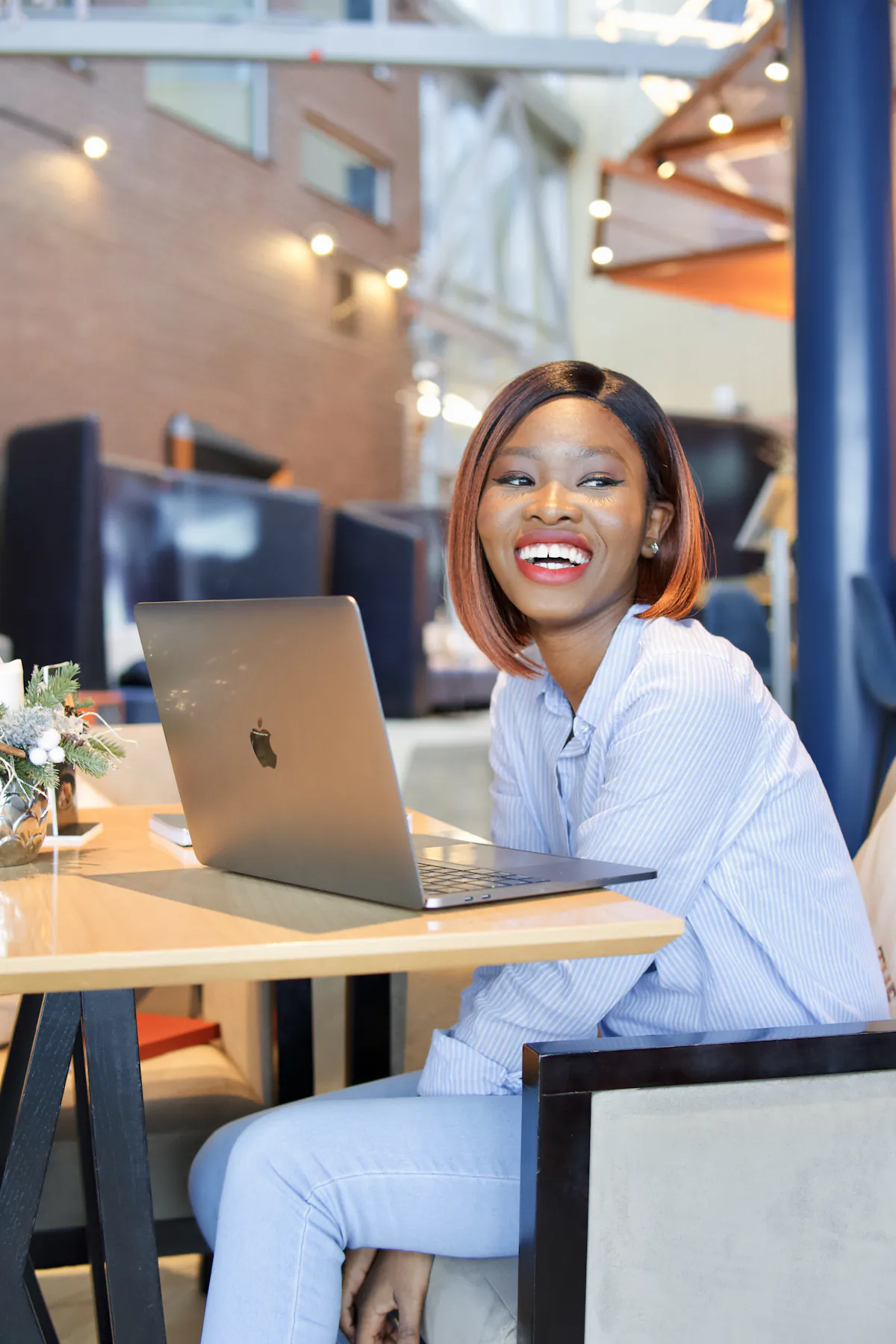 A woman sits at her laptop in a bright café, laughing and turning away from the screen.
