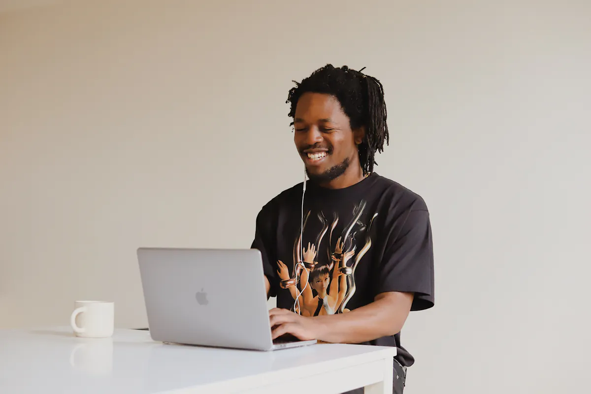 A man laughs at his laptop at a white desk, a mug beside him.