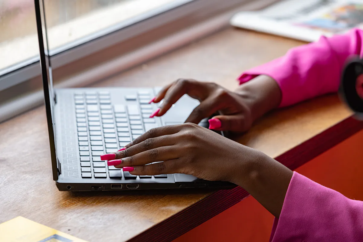 Las manos de una mujer escriben en el teclado de una laptop junto a una ventana.