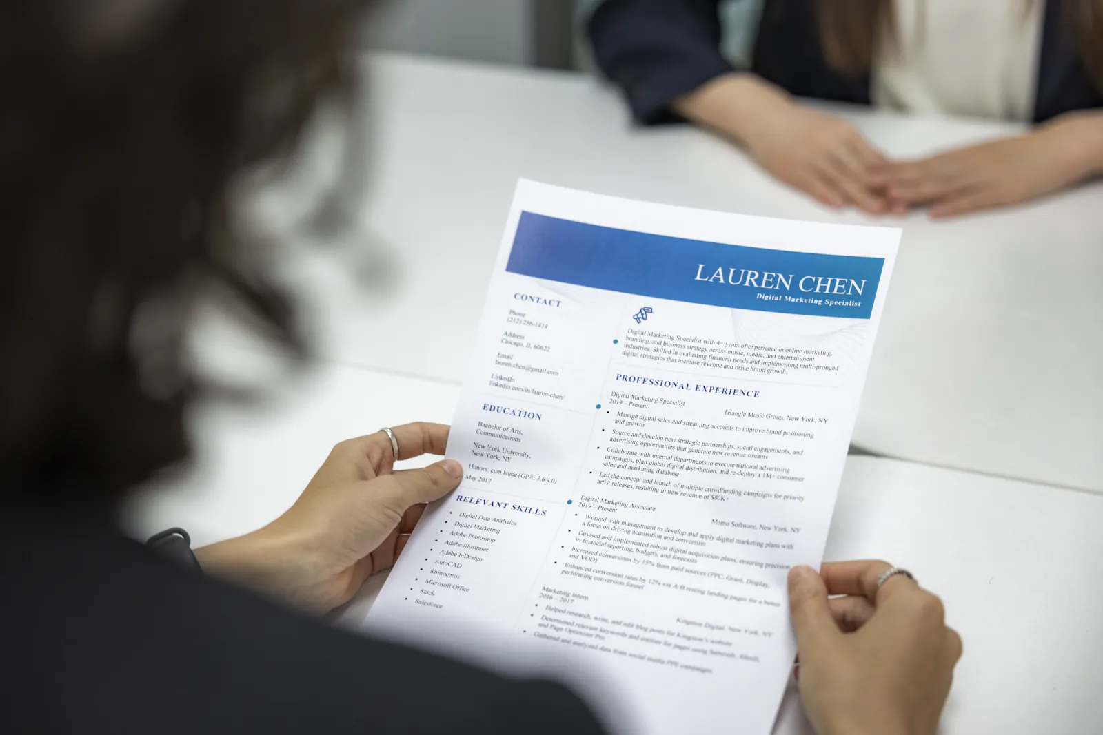 A person holds a printed résumé across a white table while another person's hands rest opposite them.