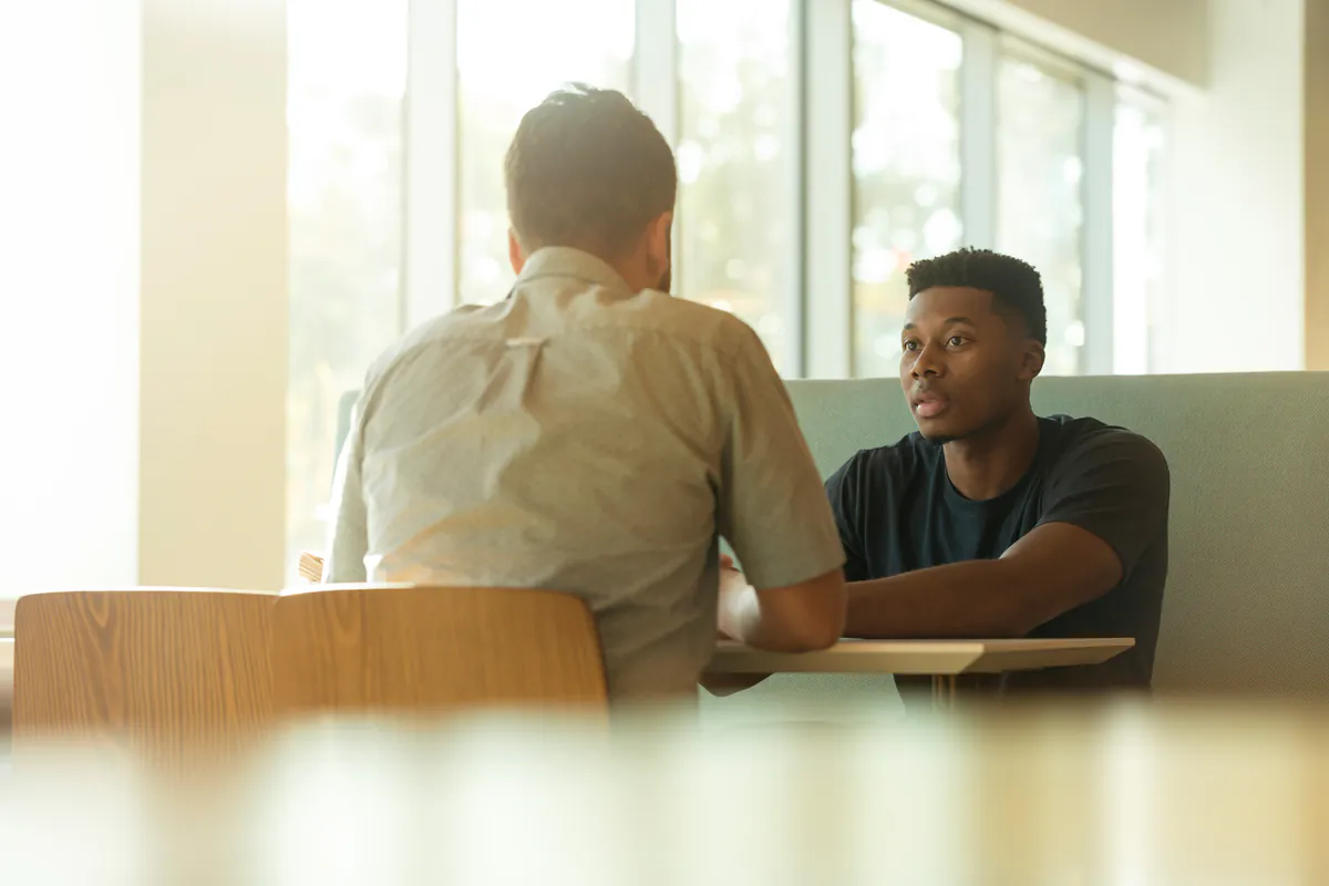 Two men talk across a table in a room full of daylight, one seen from behind.