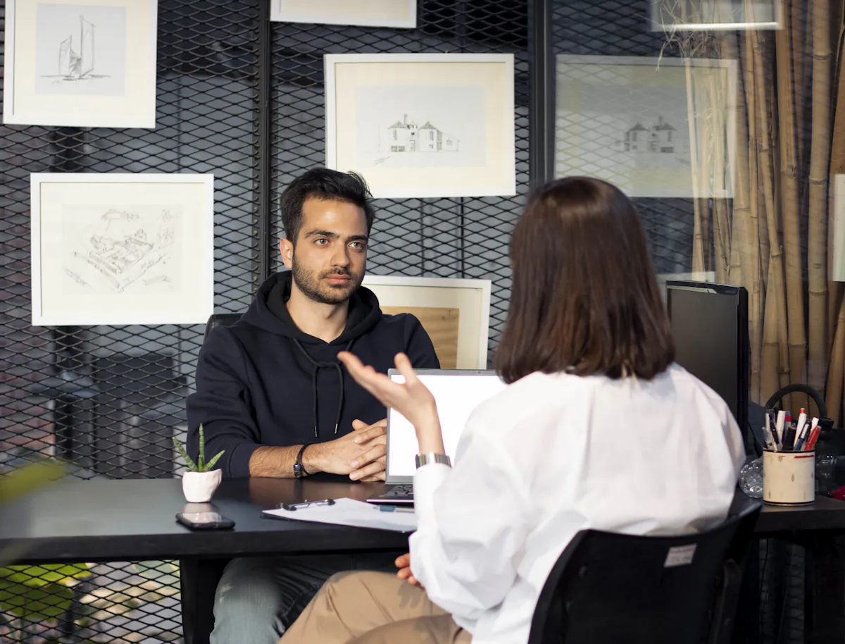 A man talks across a desk to a woman seated opposite him in a small office.