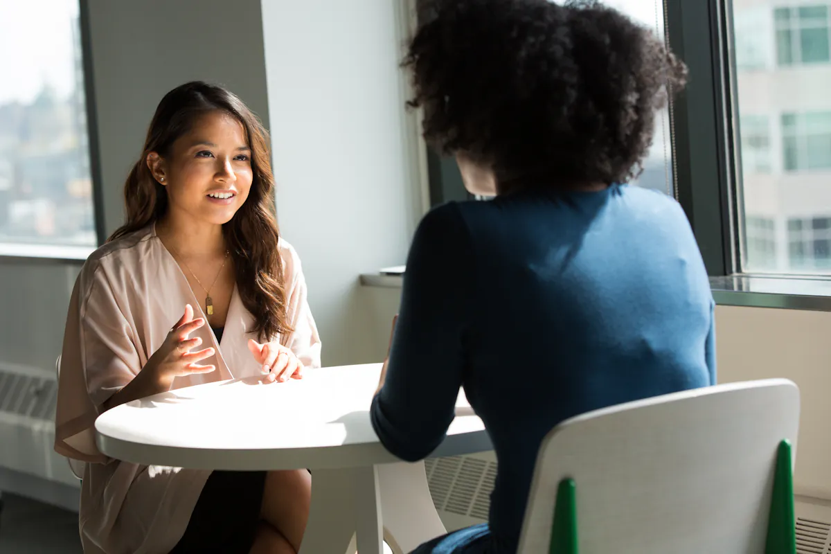 Two women talk across a small round table in front of a window overlooking a city.